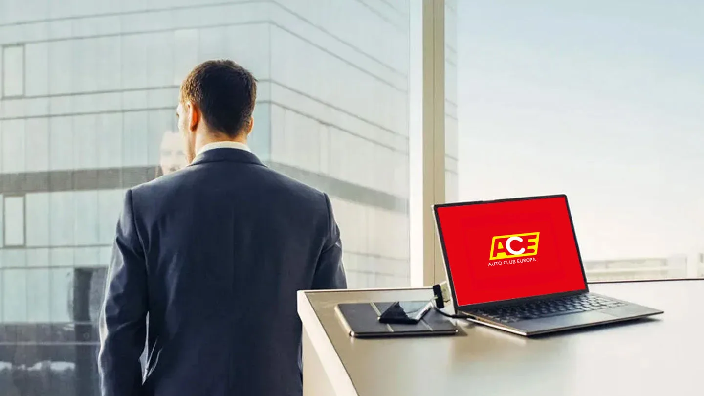 Mann im Anzug steht am Fenster mit Rücken zum Raum und auf dem Schreibtisch steht ein Laptop mit ACE Logo